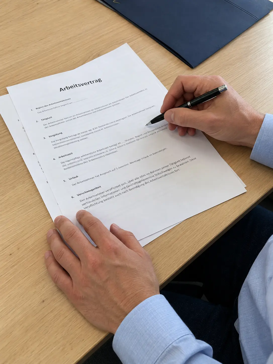 Hand unterschreibt einen Arbeitsvertrag mit dem Kugelschreiber am Schreibtisch, daneben eine blaue Mappe
